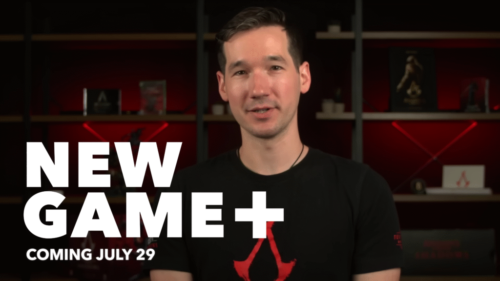 A developer from Ubisoft stands in front of a red-lit shelf wearing an Assassin’s Creed shirt, announcing the New Game+ mode with bold white text on the screen that reads: “NEW GAME+ COMING JULY 29.” The setting appears to be a studio or developer update video.