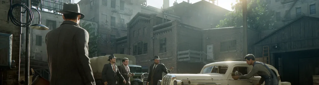 The Complete Mafia Game Story Timeline Explained | 1920 - 1968 3 A scene from Mafia 1 showing Tommy Angelo in a gray overcoat and fedora entering a back alley garage filled with fellow mobsters. Classic 1930s cars are parked nearby, while a mechanic works on one of them. The setting is industrial, with brick buildings and fire escapes under the golden afternoon light—capturing the gritty atmosphere of Lost Heaven.